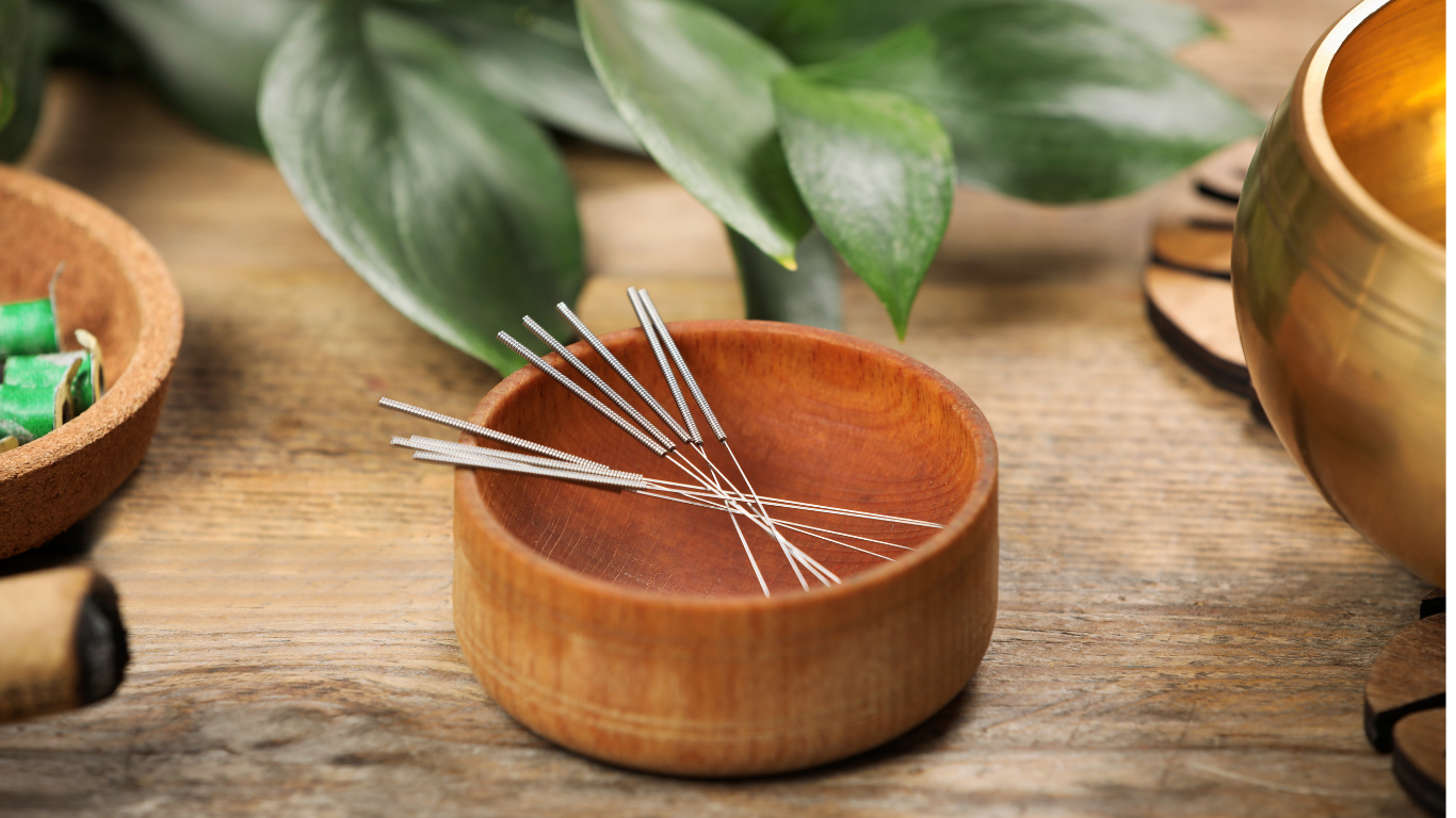 Wooden bowl of acupuncture needles on a rustic table with green leaves and a brass bowl nearby