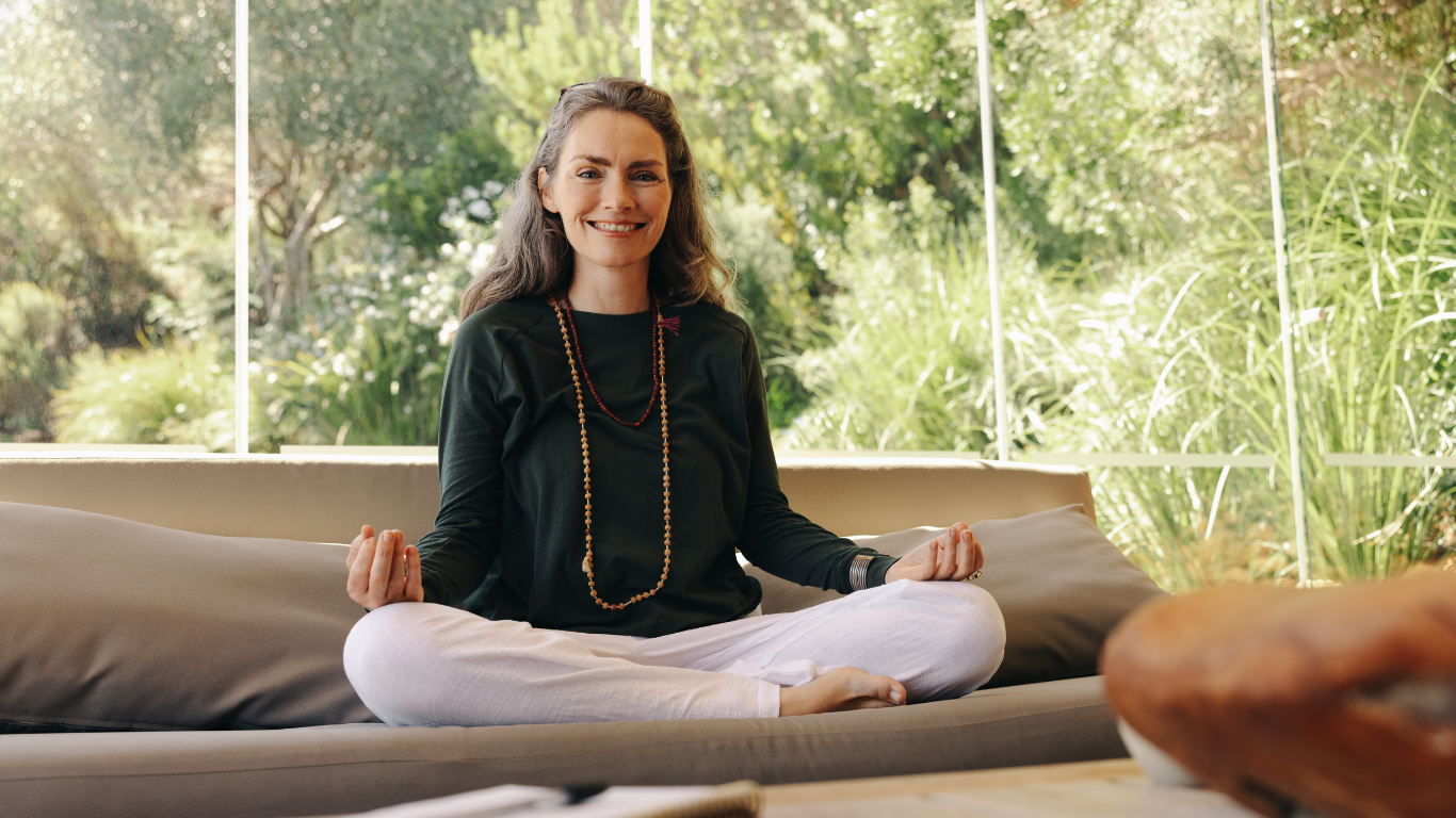 Person meditating cross-legged on a sofa in a bright room with large windows and greenery outside