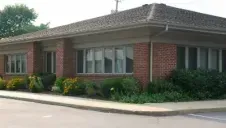 Brick building with a dark roof, large windows, and trimmed shrubs beside a paved street.