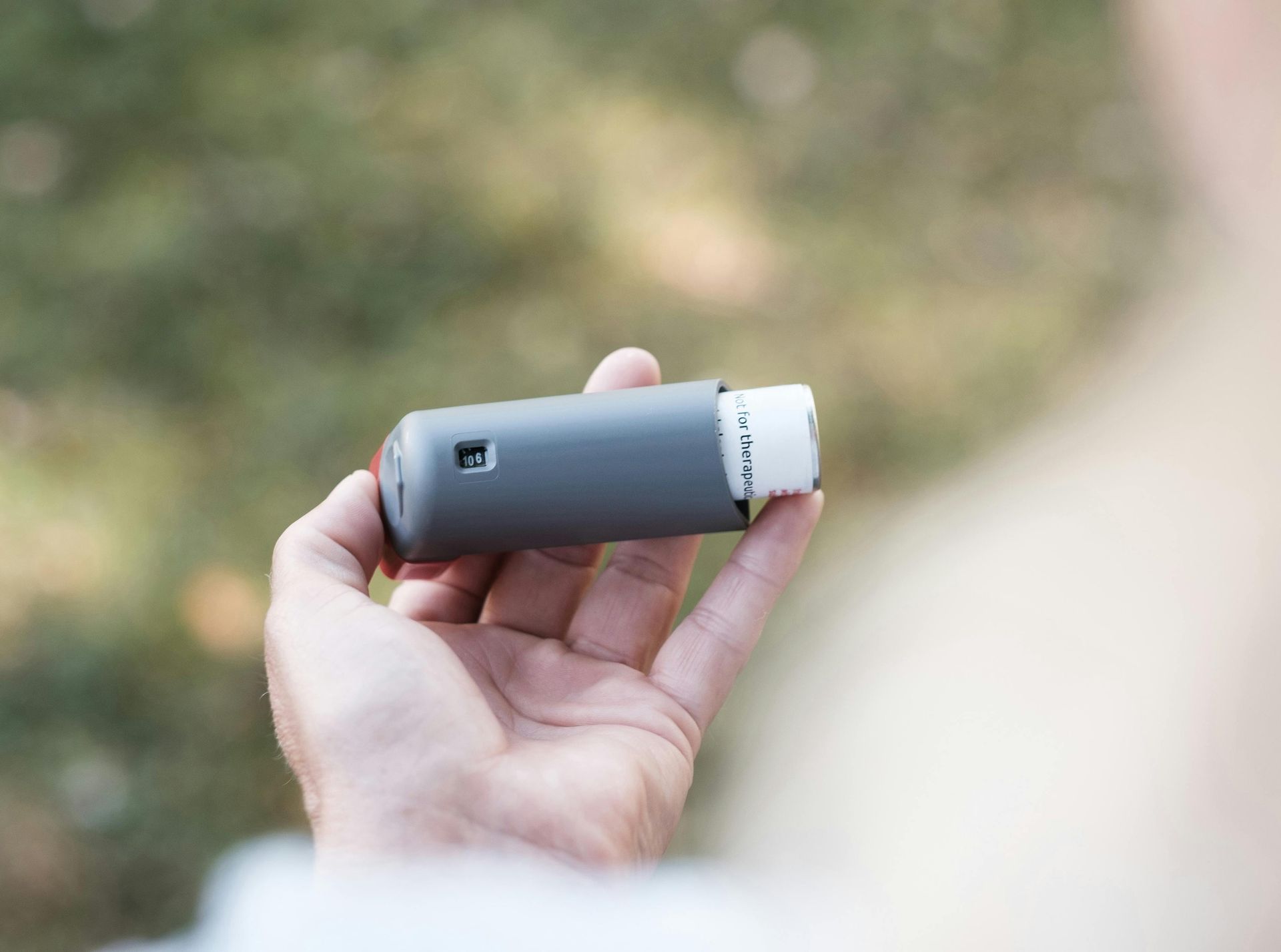 Hand holding a gray inhaler against a blurred outdoor background
