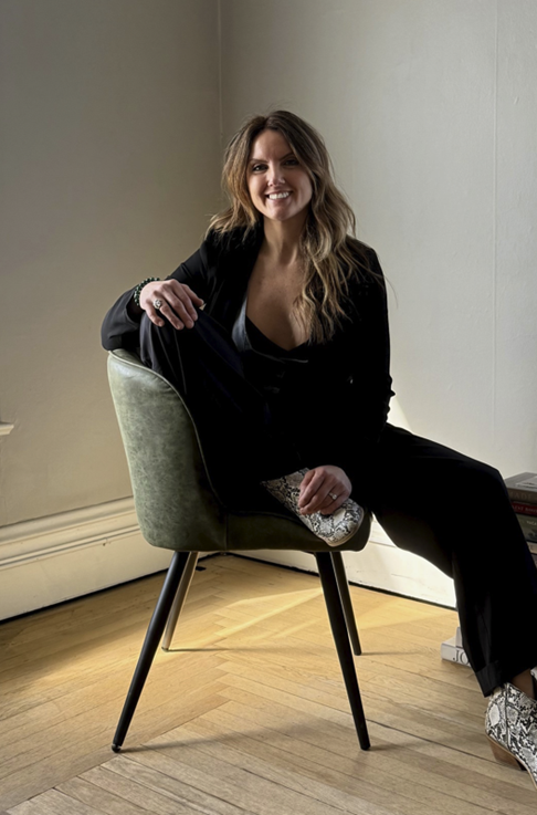 Woman in black suit, seated, smiling by windows.