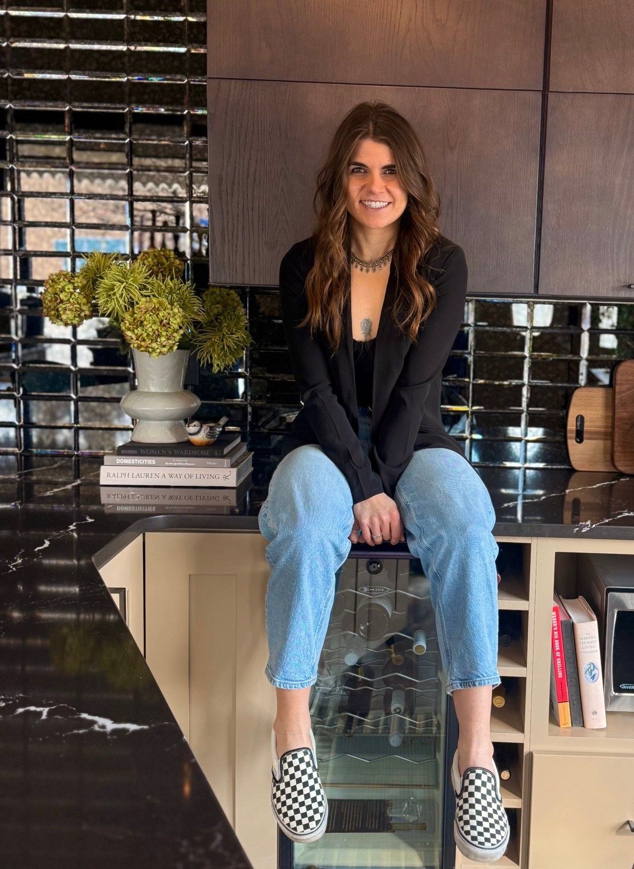Woman sitting on a kitchen counter, wearing a black blazer, jeans, and checkerboard shoes.