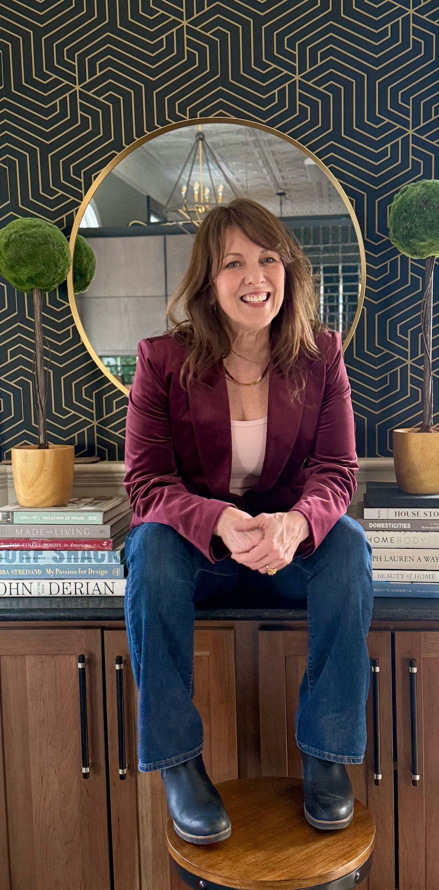 Woman smiling, seated on a stool. Wearing a burgundy blazer and jeans, in front of a mirror and wall décor.