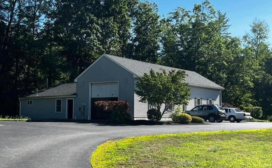 White industrial building with open garage door, parked pickup truck, and trees along a paved driveway | Cota's Auto Repair