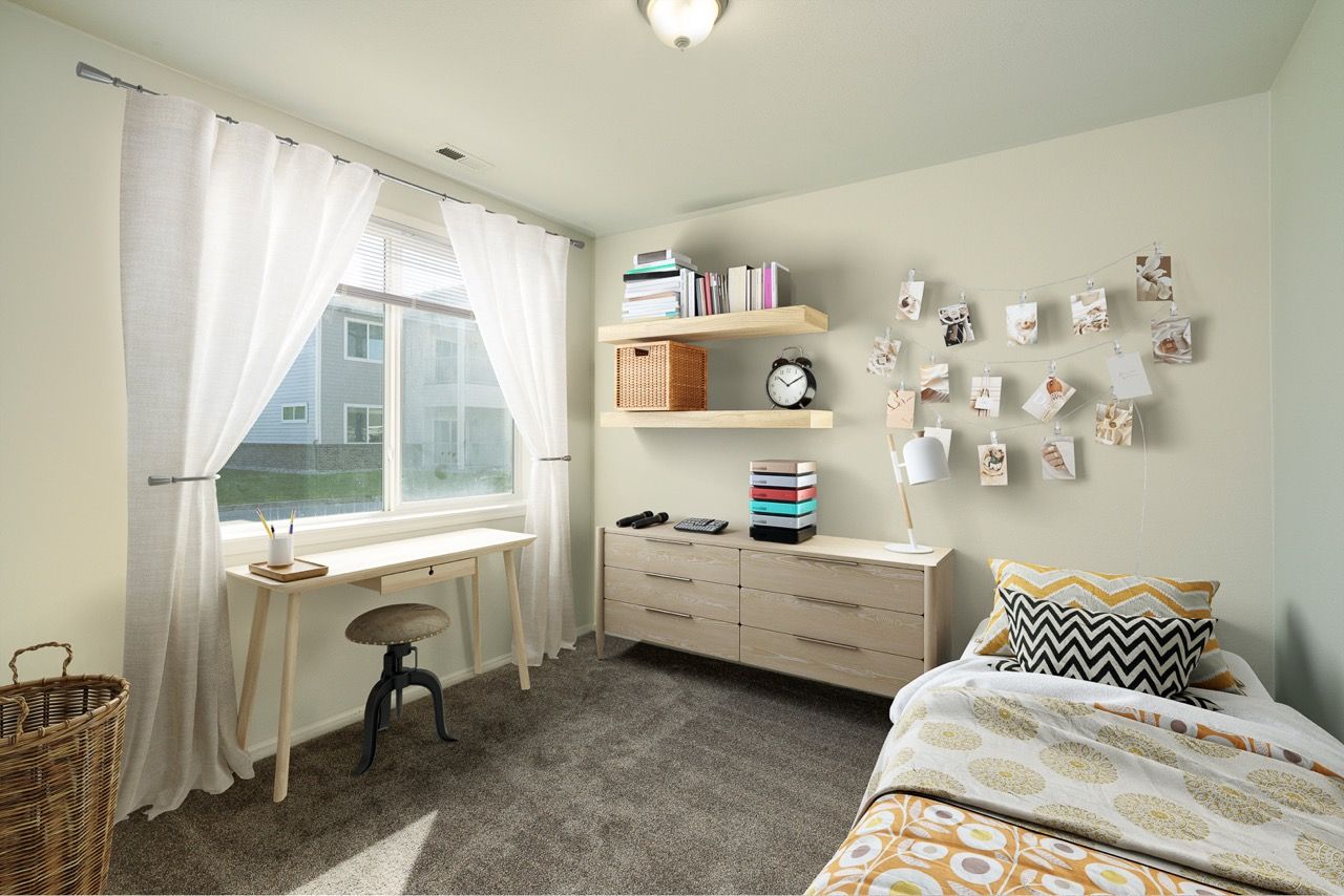 Sunlit bedroom with a window desk, light wood dresser, shelves, and a wall photo garland.