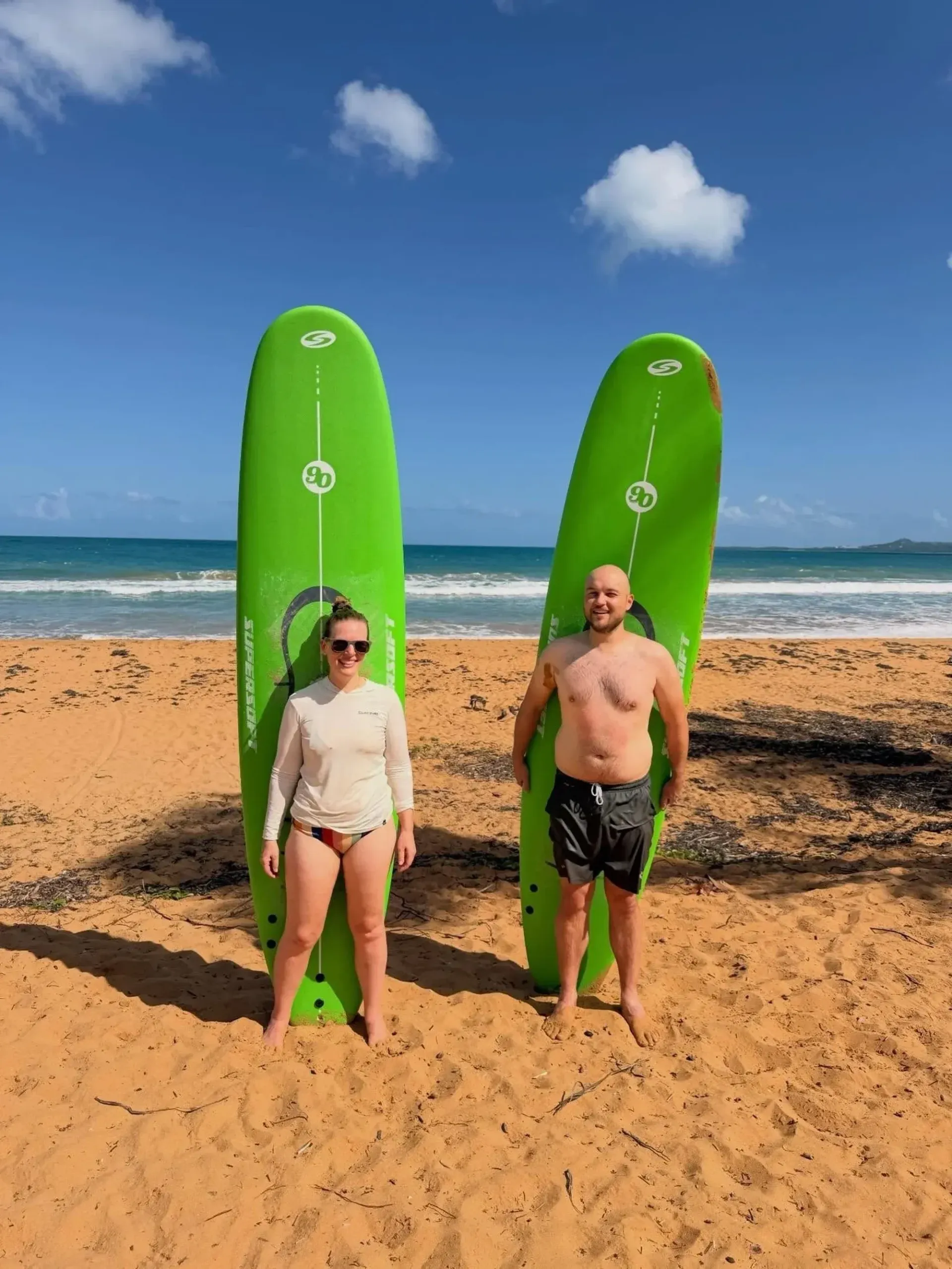 Two people holding green surfboards on a sunny beach with ocean and blue sky in the background
