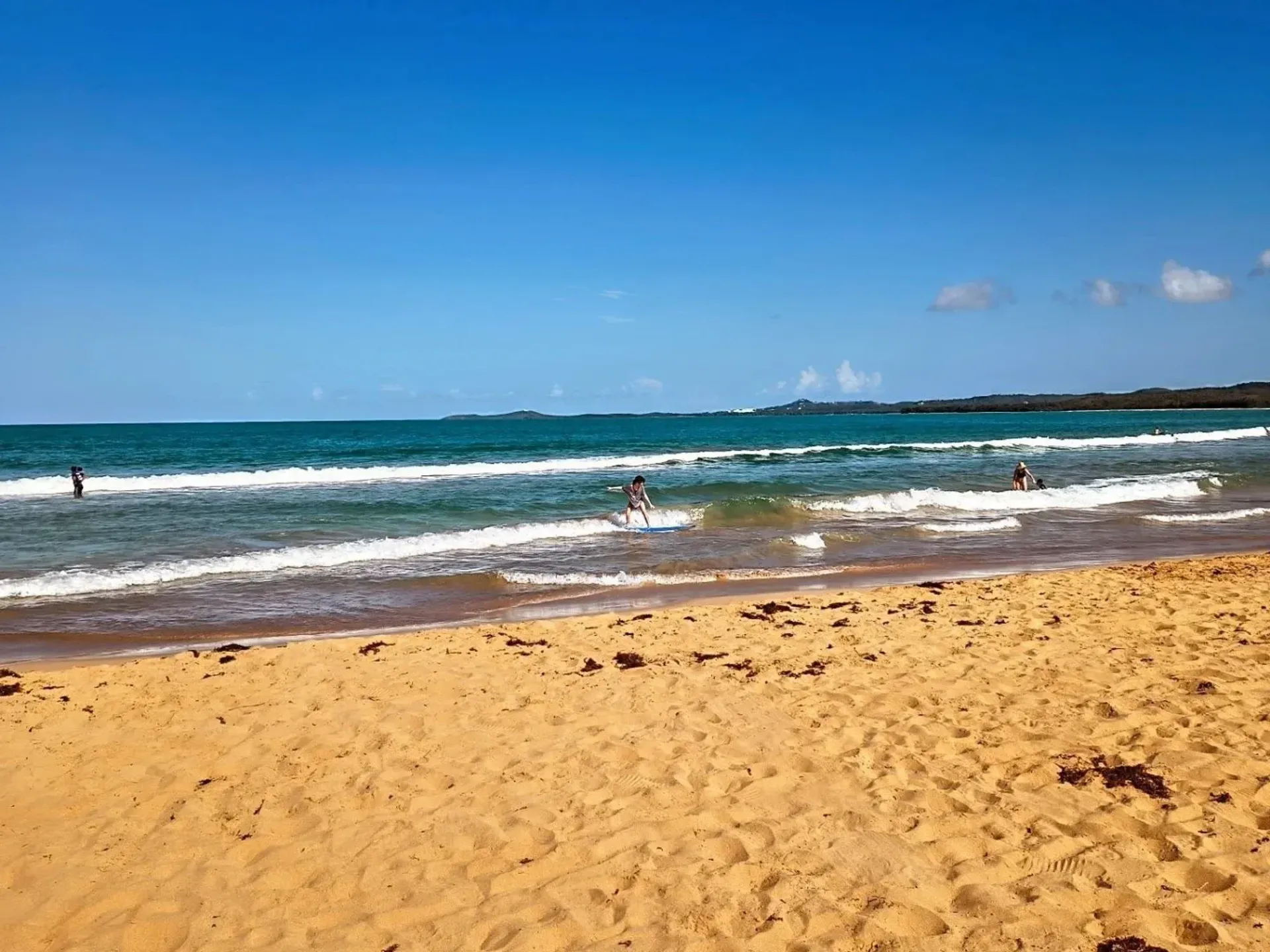 Sunny beach with golden sand, blue ocean waves, and a few people swimming near shore