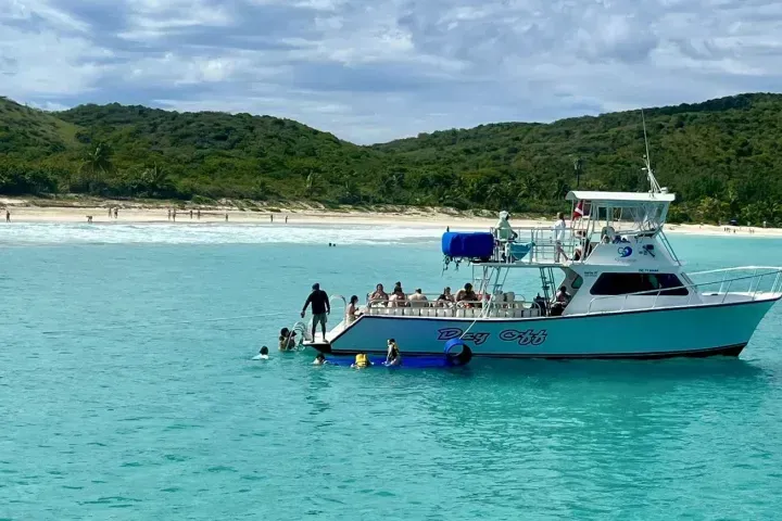 White boat with people floating in turquoise water near a tropical beach and green hills