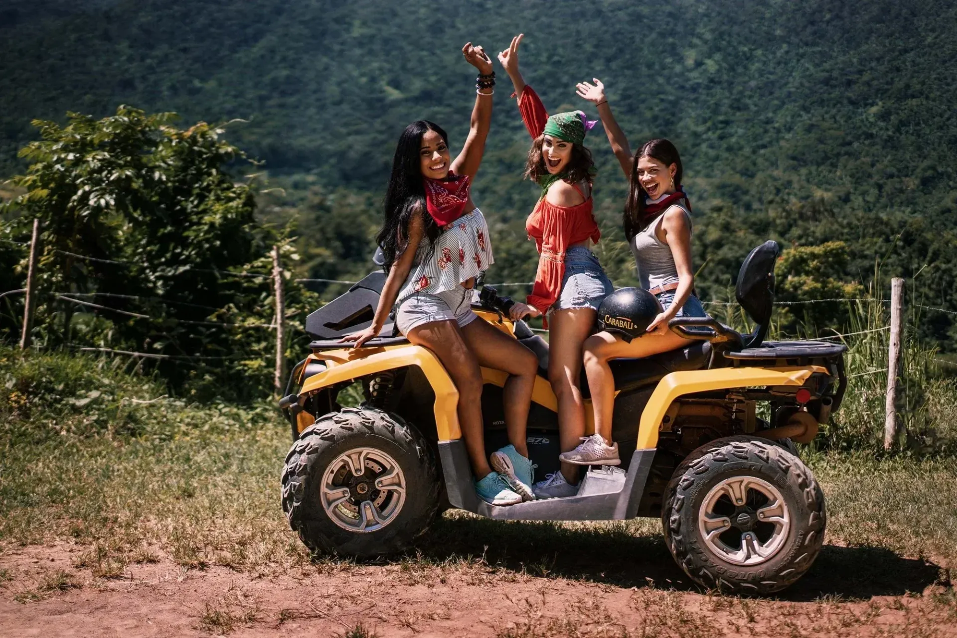 Four friends posing on a yellow ATV in a muddy field, with green hills in the background.
