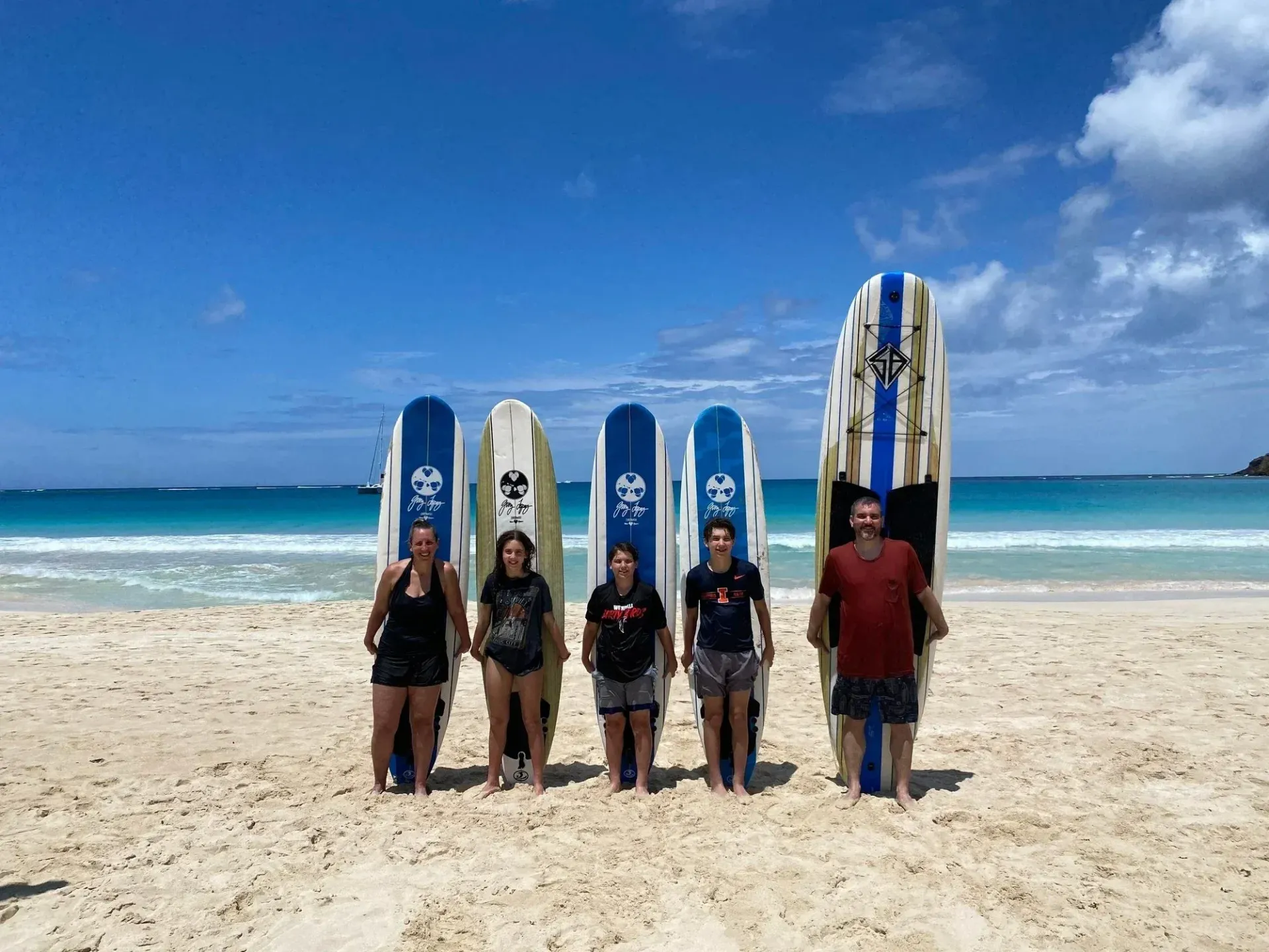 Five people standing on a sunny beach with surfboards, ocean and blue sky behind them