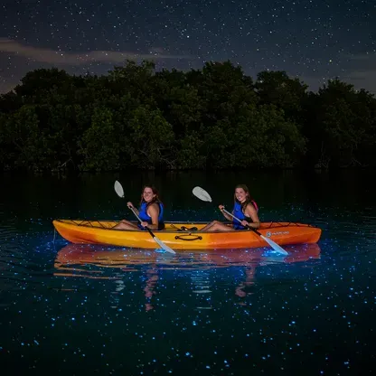 Two kayakers in an orange canoe on glowing blue water at night under a dark sky