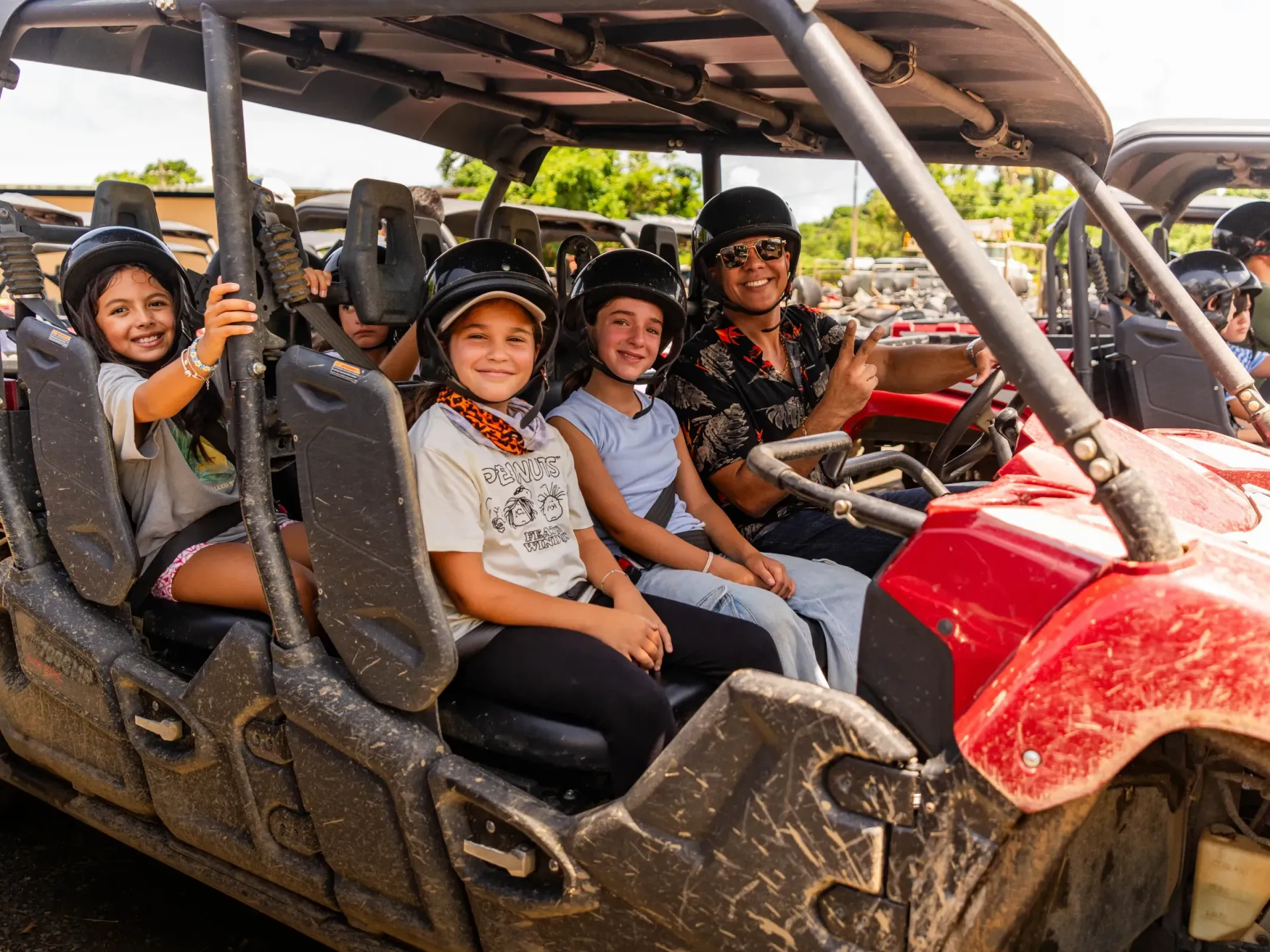 Four people smiling in a muddy off-road buggy, with helmets and a red front hood.