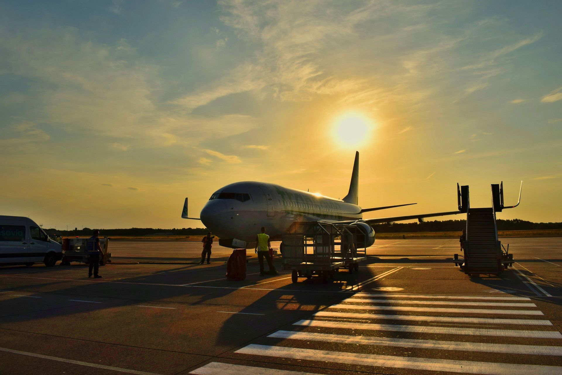 Passenger jet on runway at sunset, with ground crew and vehicles silhouetted against the orange sky