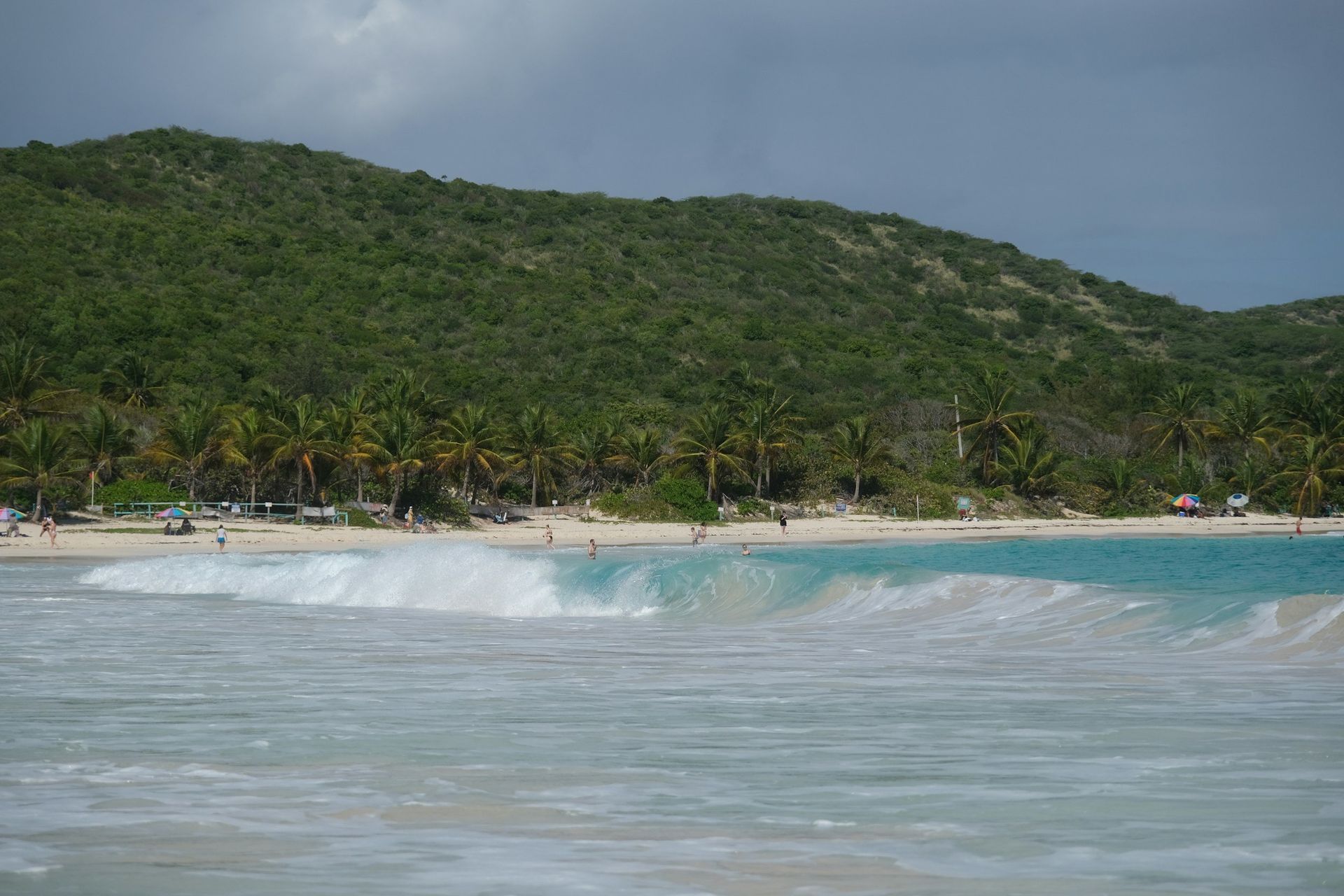 Tropical beach with turquoise waves, sandy shore, and lush green hillside under cloudy sky