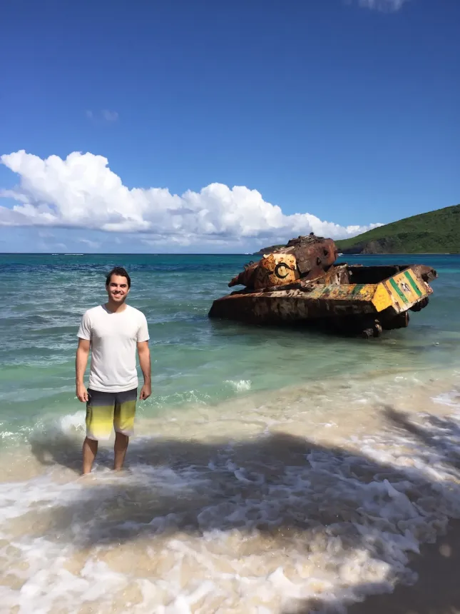 Person standing in shallow tropical water beside a rusted tank near the shore