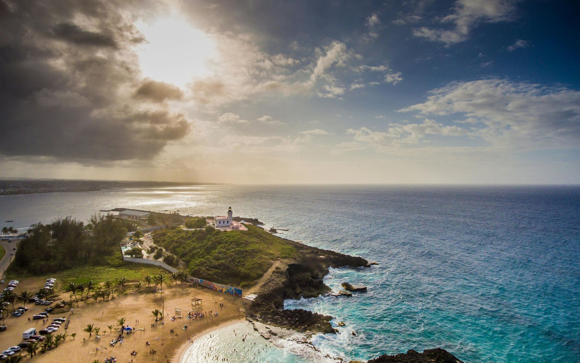 Sunlit rocky coastline with turquoise water, beachgoers, and a cloudy sky at sunset