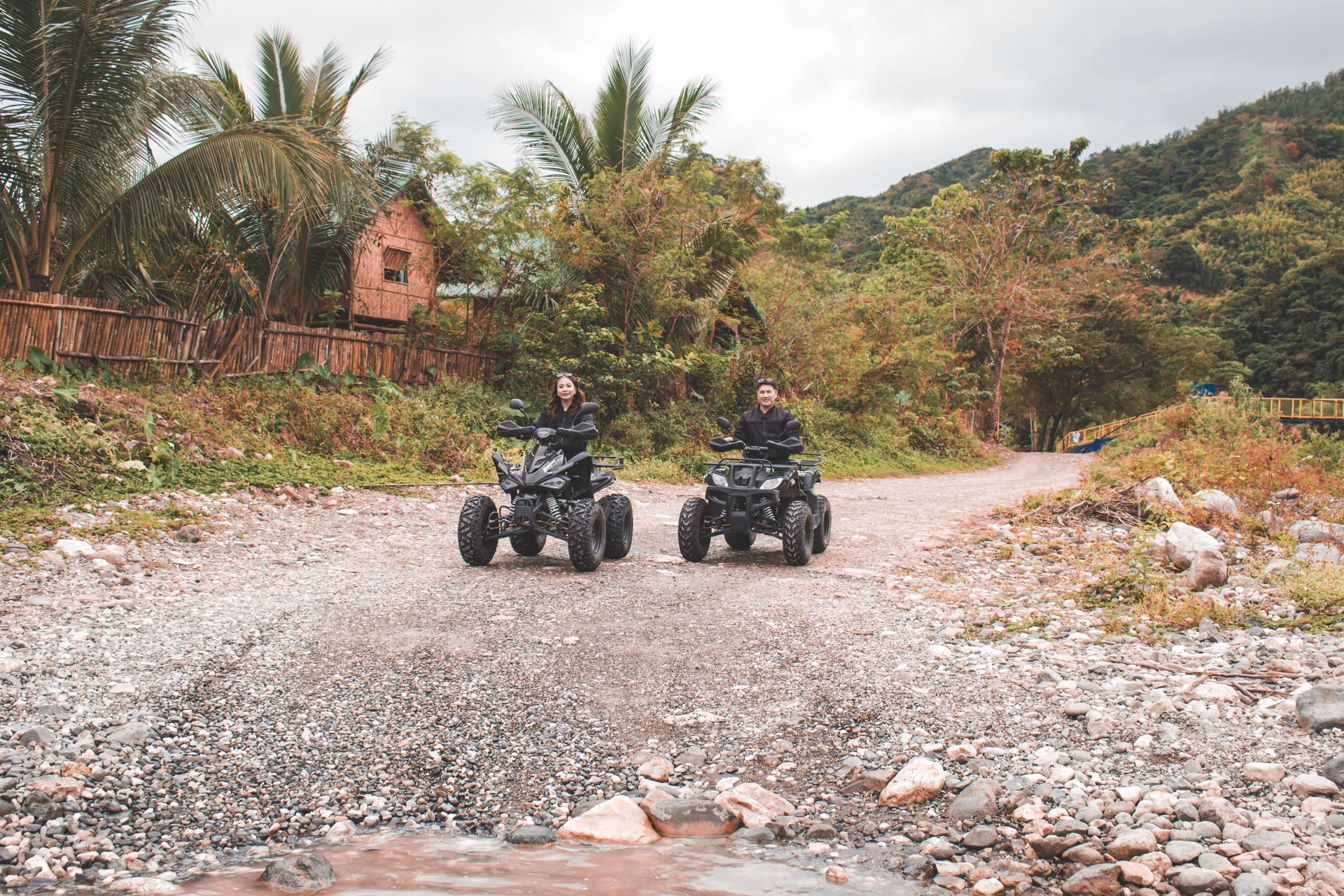 Two ATV riders on a rocky dirt road with palm trees and hills in the background