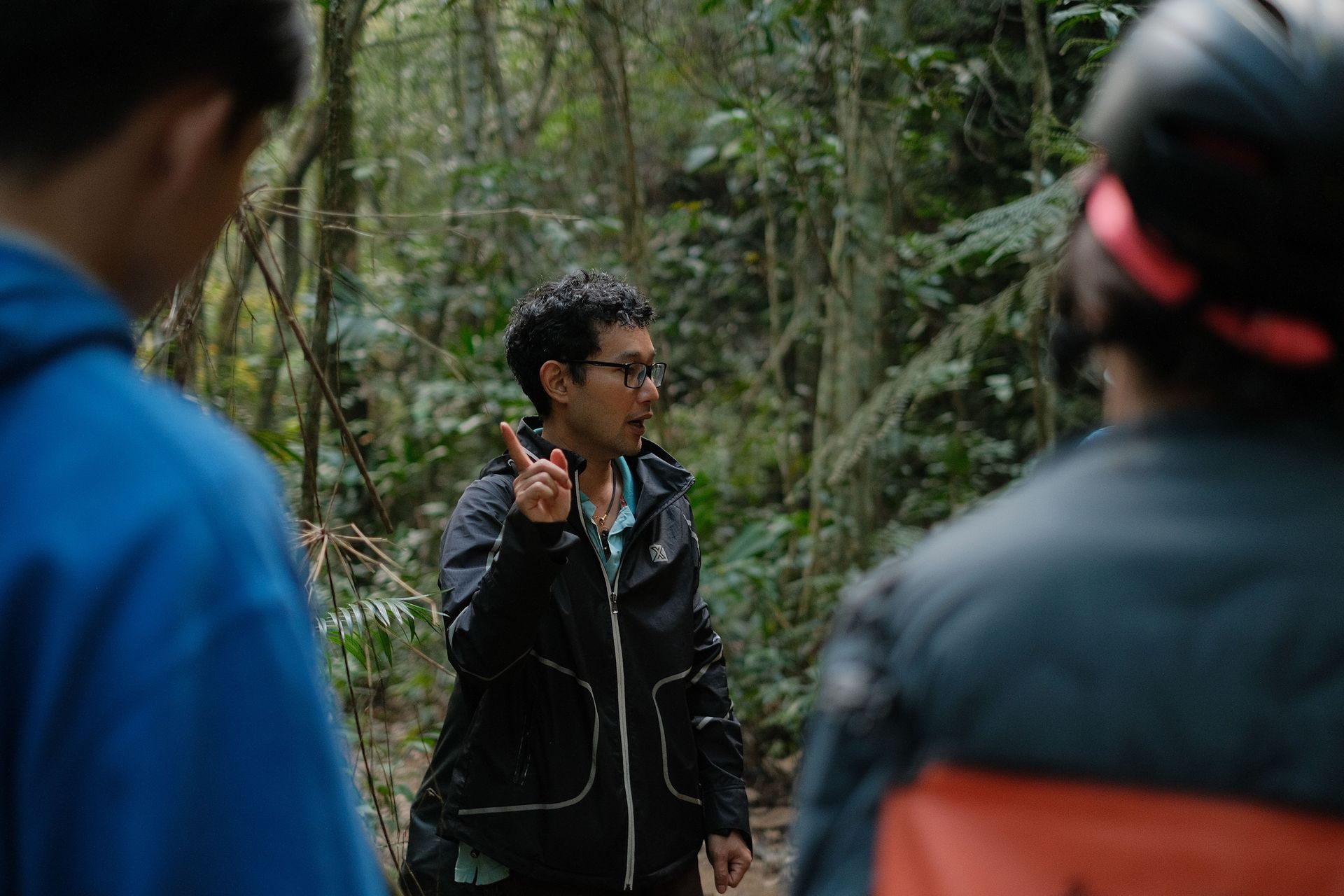 Person in a black jacket gesturing in a wooded area, seen between two blurred people in the foreground.