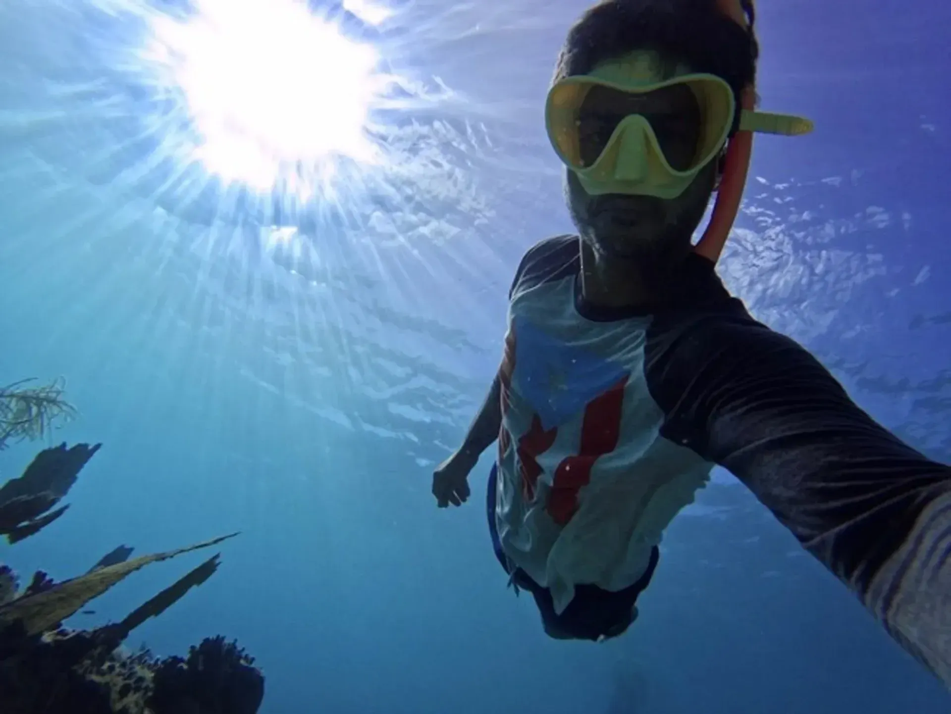 Snorkeler underwater taking a selfie, wearing a green mask and sunlit blue ocean around them