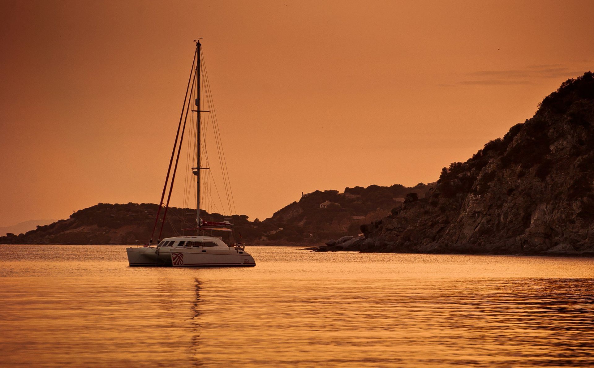 Sailboat silhouetted on calm orange water at sunset near a rocky coastline