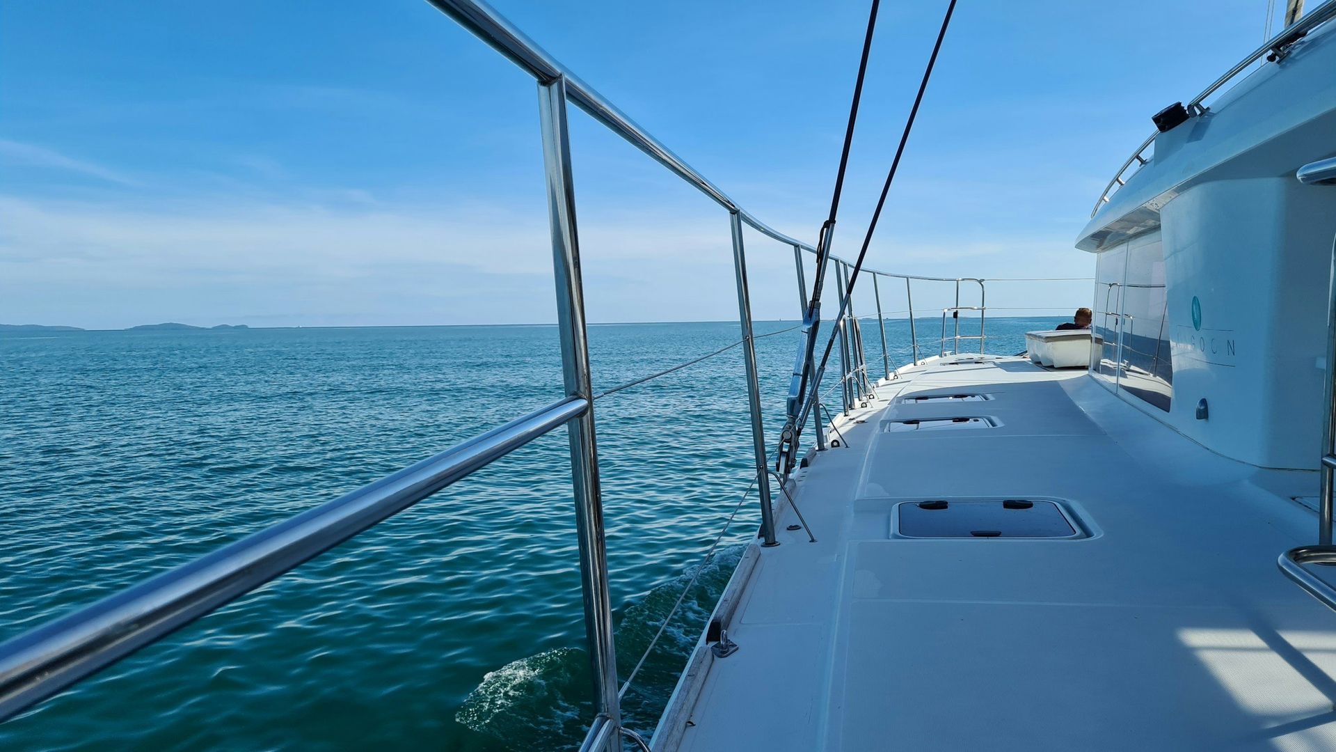 Sailboat deck on calm blue water with railings and distant shoreline under a clear sky