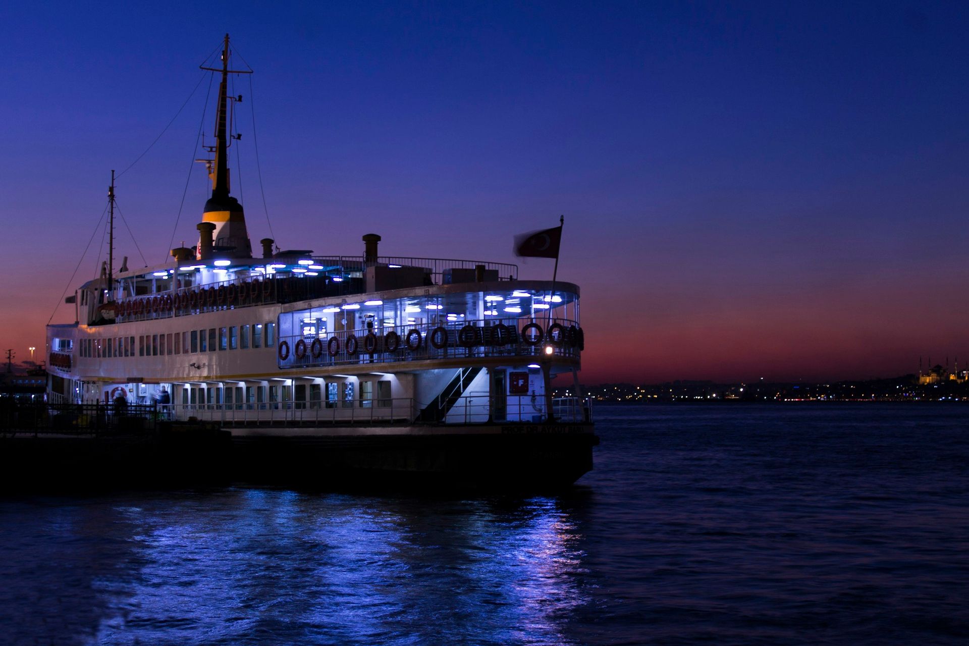 Illuminated ferry boat on calm water at dusk with city lights in the distance