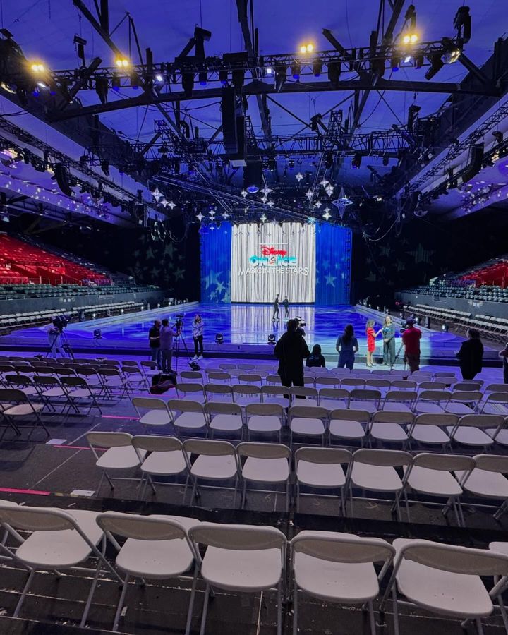 Empty arena with rows of white chairs facing a stage lit with blue, white, and red lights.