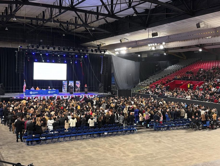 Large crowd at an event with a stage, seating, and a banner.  Many people raising their hands.