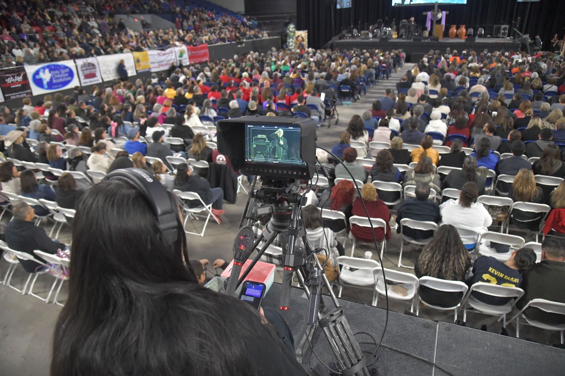 A person with headphones operates a video camera, filming an event in a large arena packed with an audience.