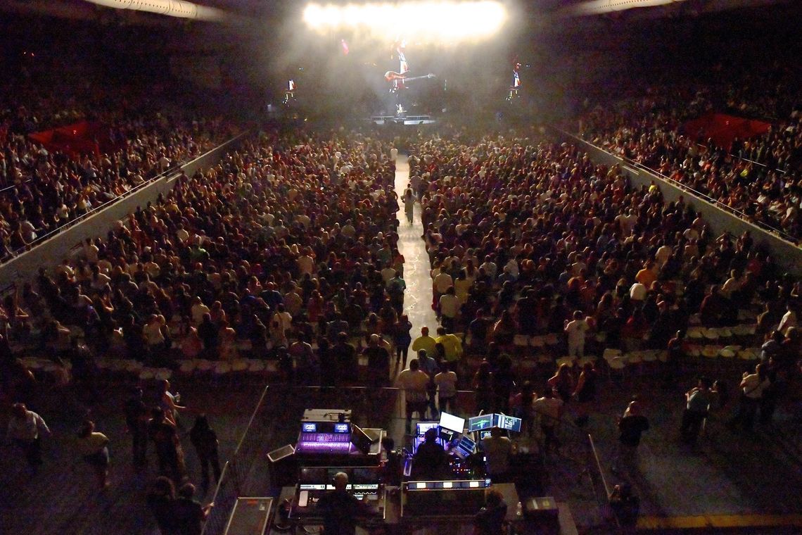 A crowded concert arena with a stage lit by bright lights. Audience members fill seats; a long path is clear.