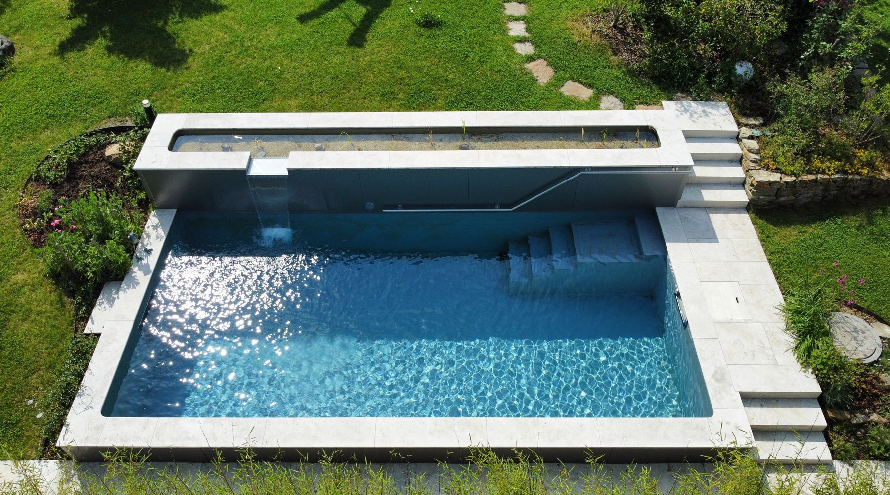 Rechteckiger Swimmingpool im Garten mit blauem Wasser, Betonterrasse und umlaufendem grünen Rasen