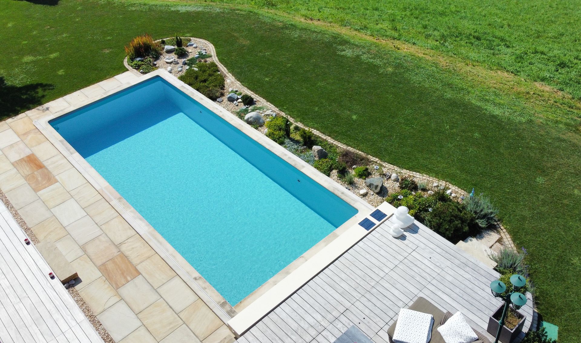 Rechteckiger blauer Swimmingpool neben einer Steinterrasse und einem grünen Rasen, von oben betrachtet.