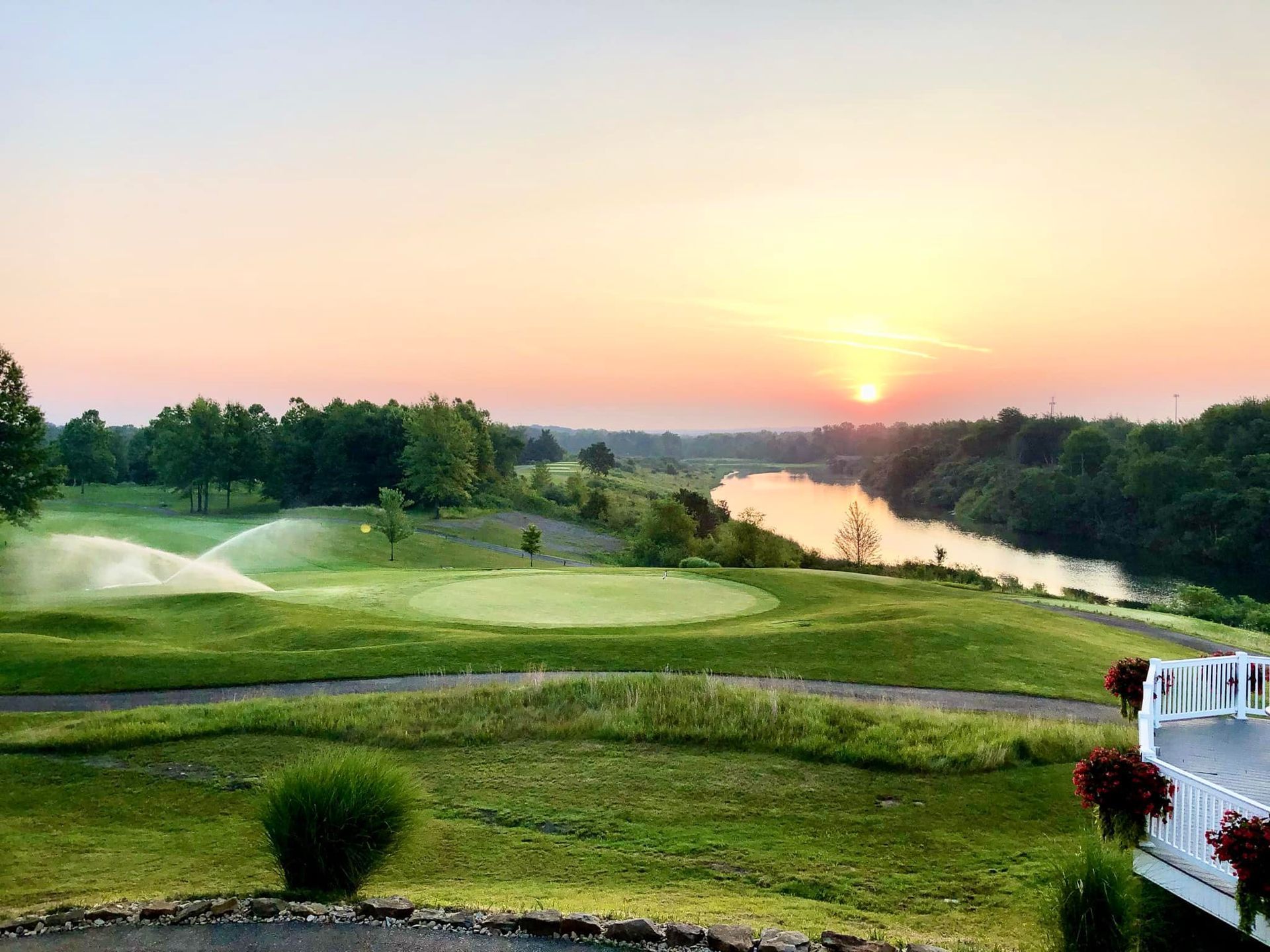 Golf course at sunset with a river and trees in the background; sprinkler watering the green.
