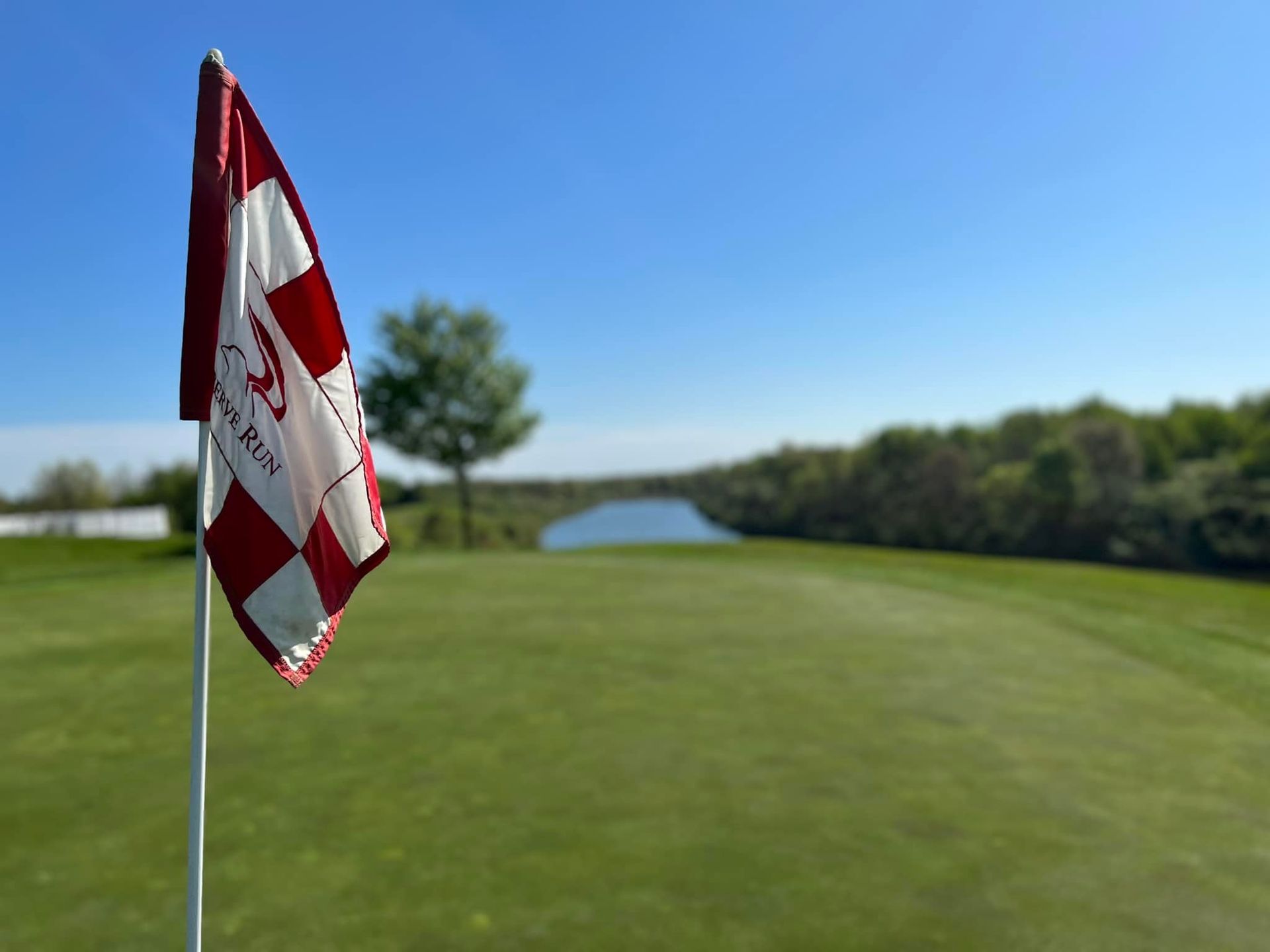 Golf course flag on green, red and white checkered, lake and trees in background under blue sky.