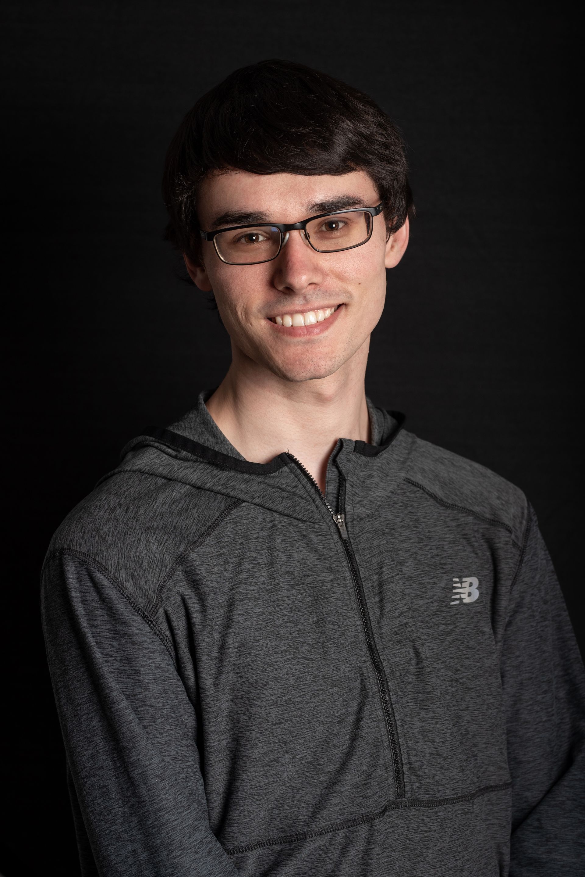 Young man wearing glasses and a gray hoodie smiles against a black backdrop.