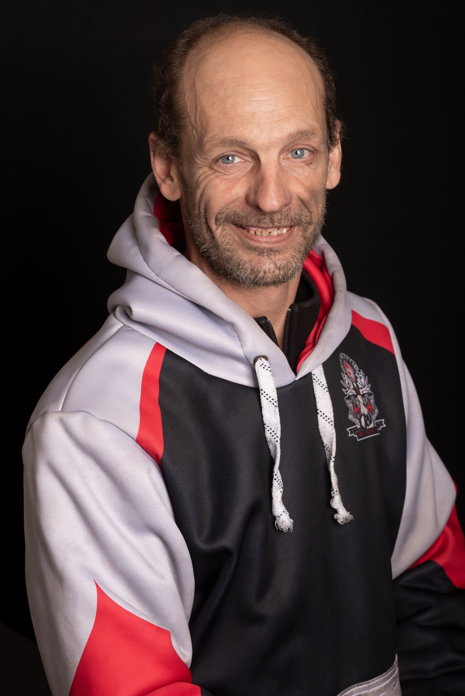 Middle-aged man with a hooded sweatshirt smiles at the camera, black background.