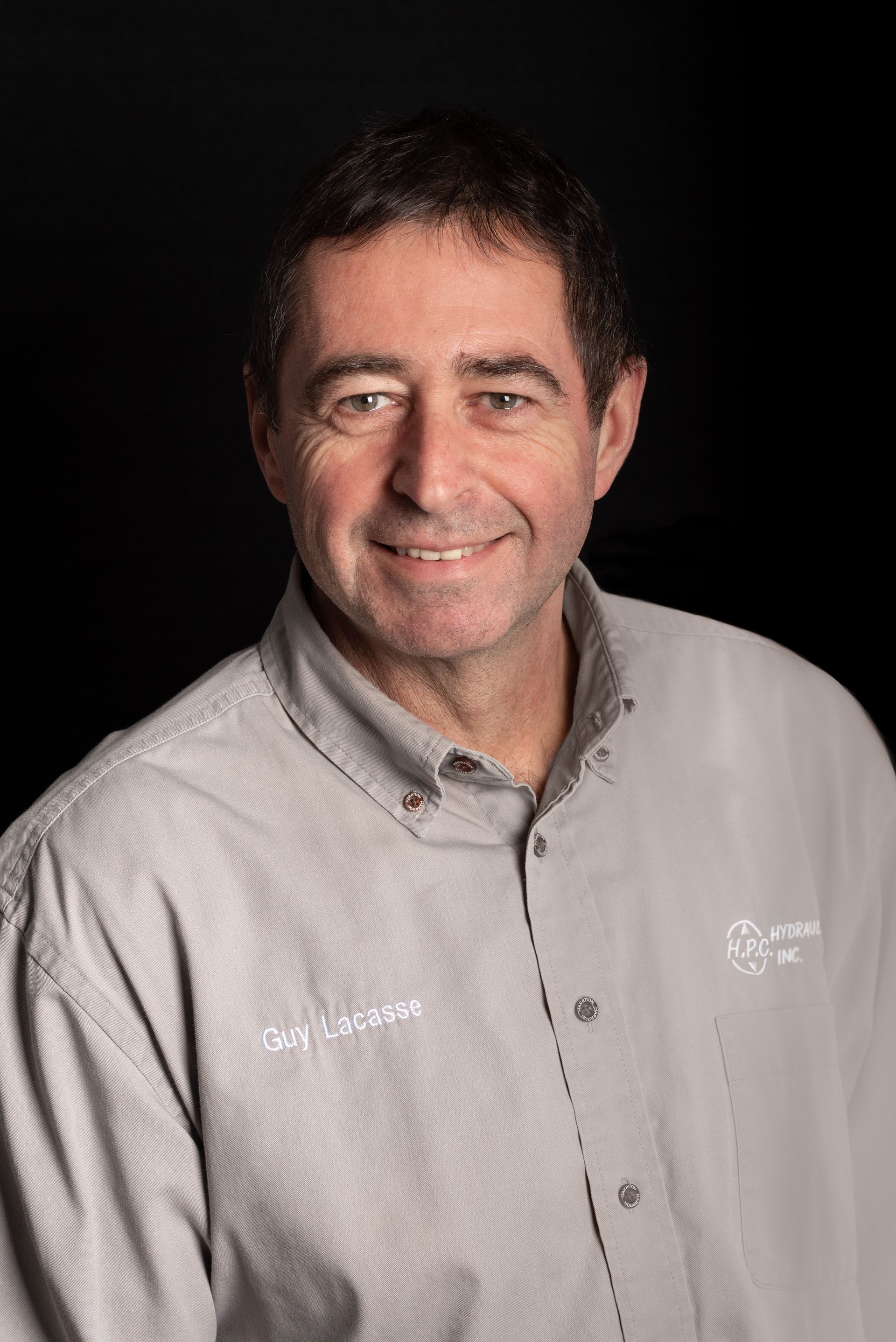 Man smiling, wearing a gray button-down shirt. Dark background.