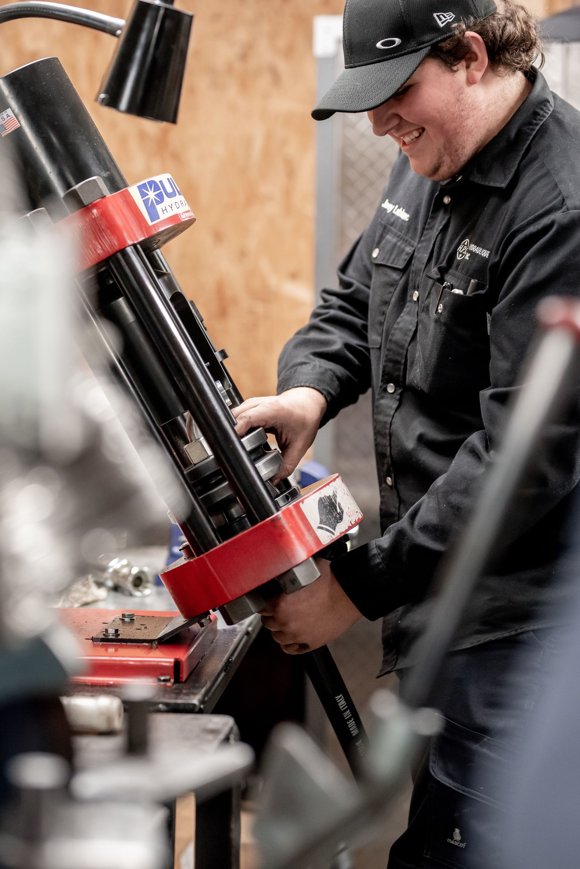 Un homme en tenue de travail noire sourit, travaillant sur une grande machine rouge et noire dans un atelier.