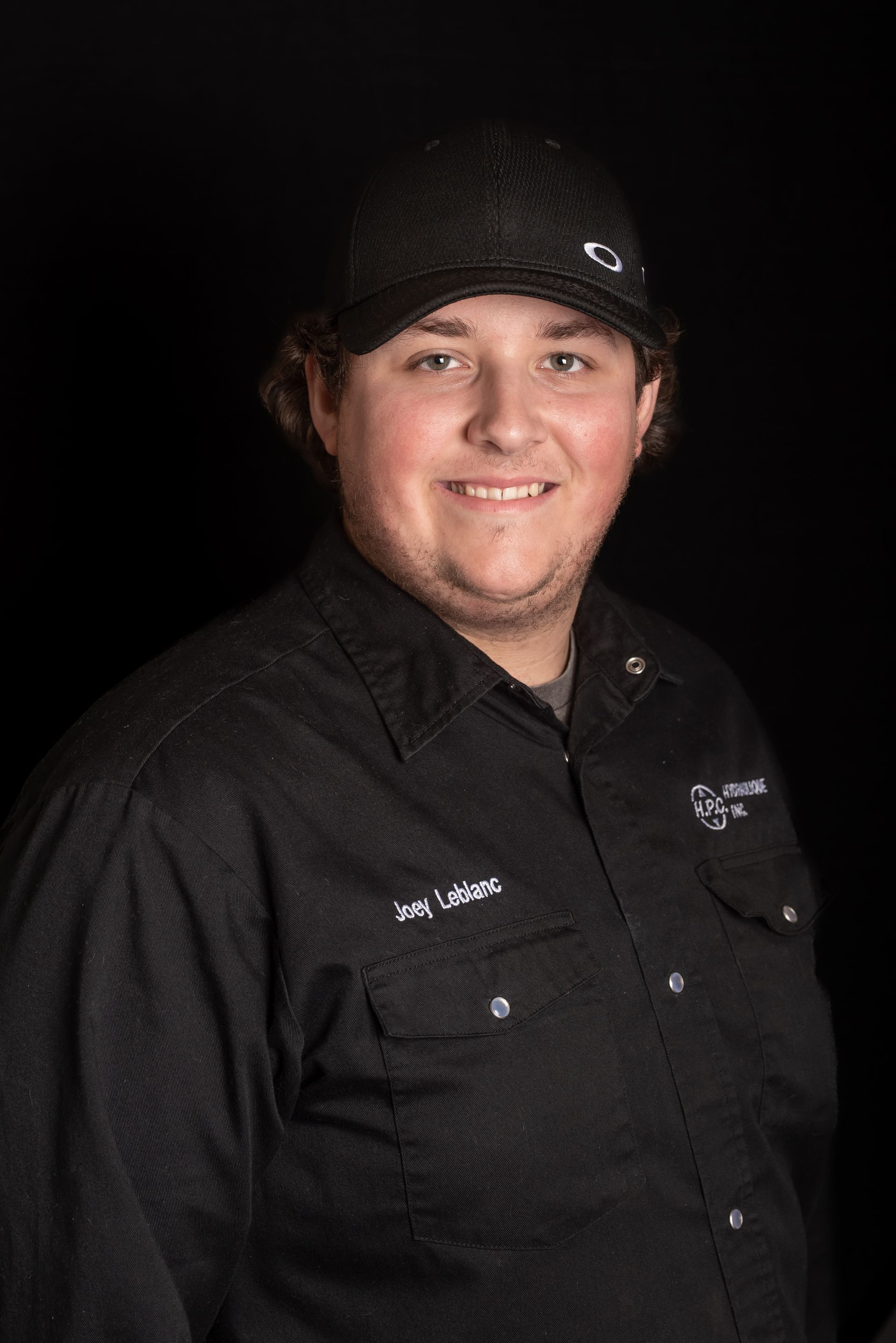 Man in black hat and shirt smiles, with a black background.