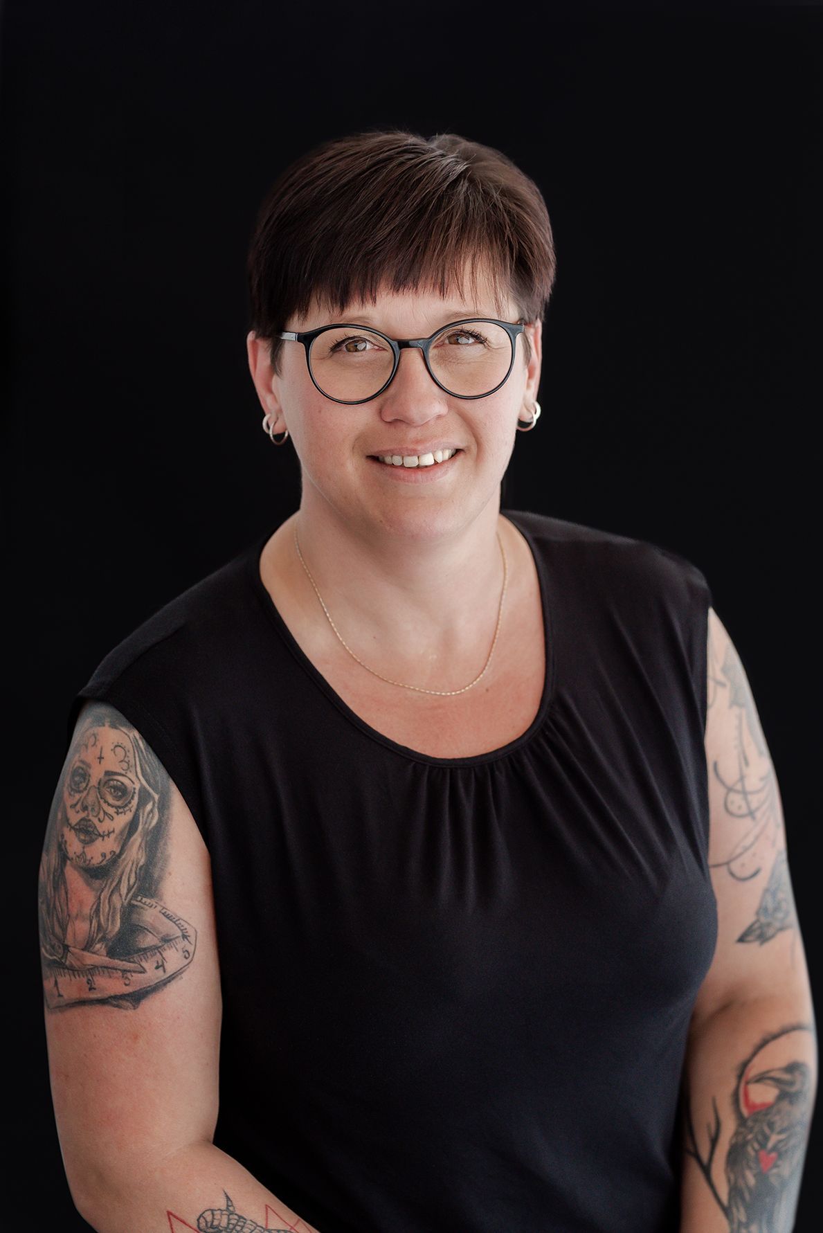 Woman with short brown hair, glasses, and arm tattoos smiles in front of a black backdrop.