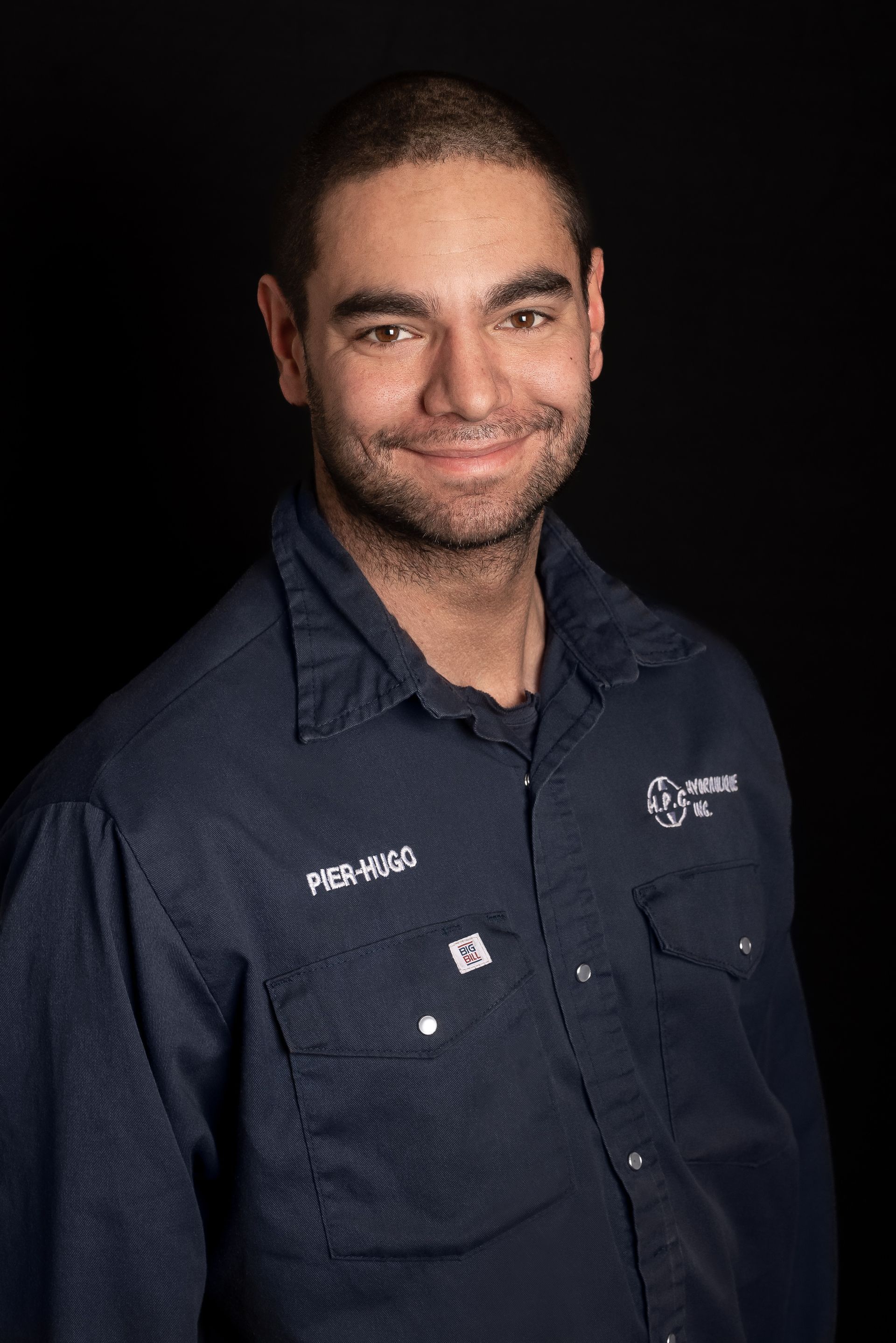 Man in a blue work shirt smiles at the camera against a black backdrop.