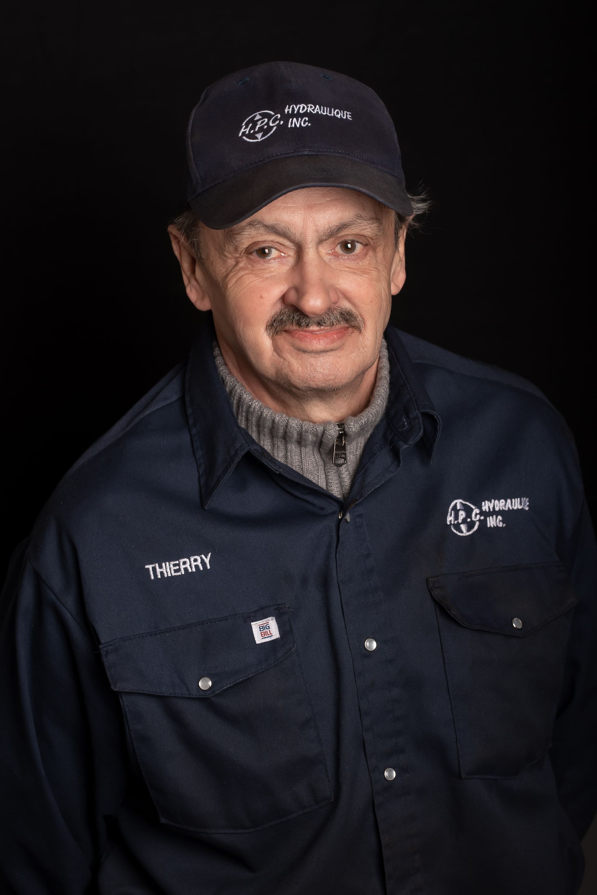 Man with mustache wearing a hat and work shirt, standing against a black backdrop.