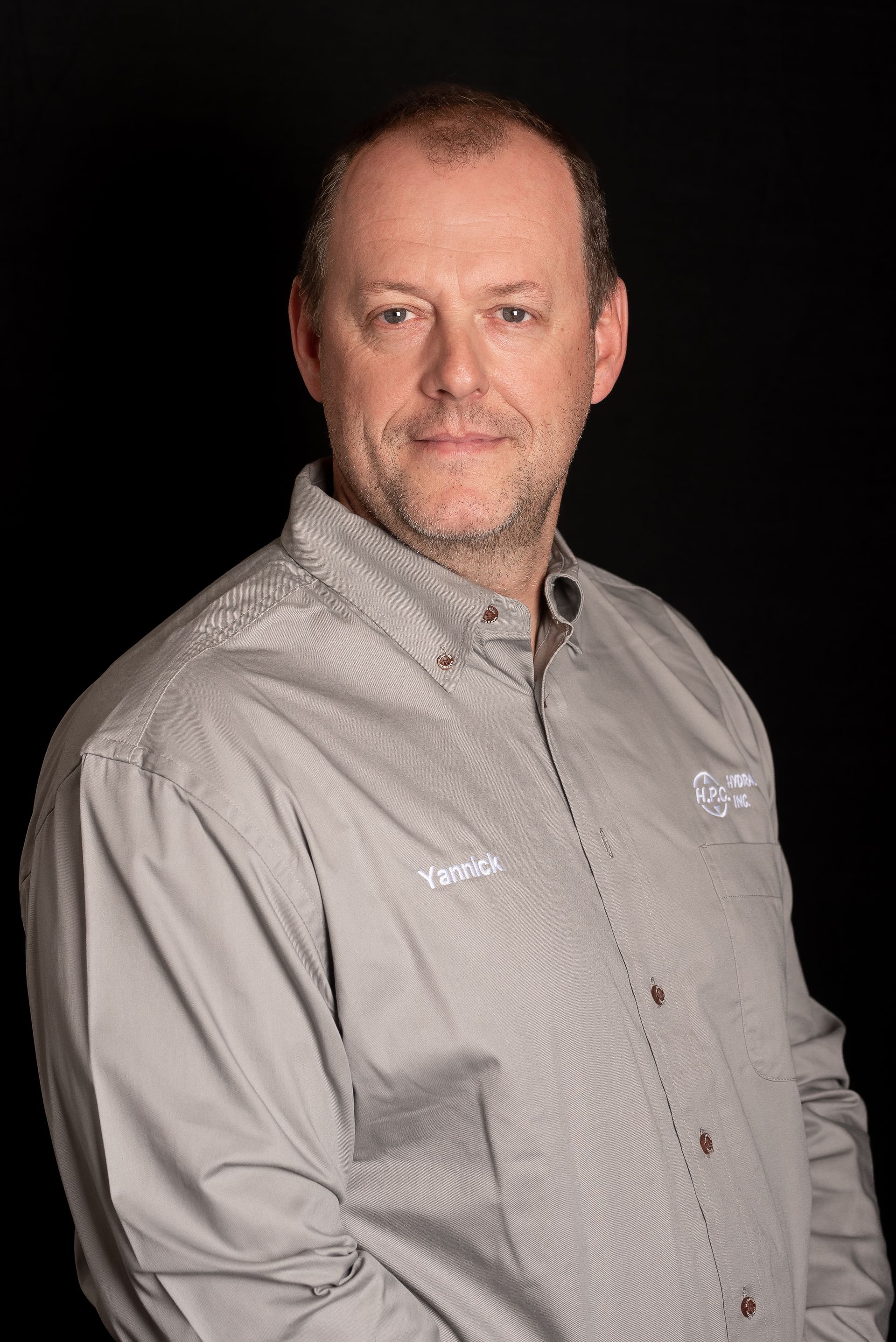 Man in gray collared shirt, slightly smiling against a black background.