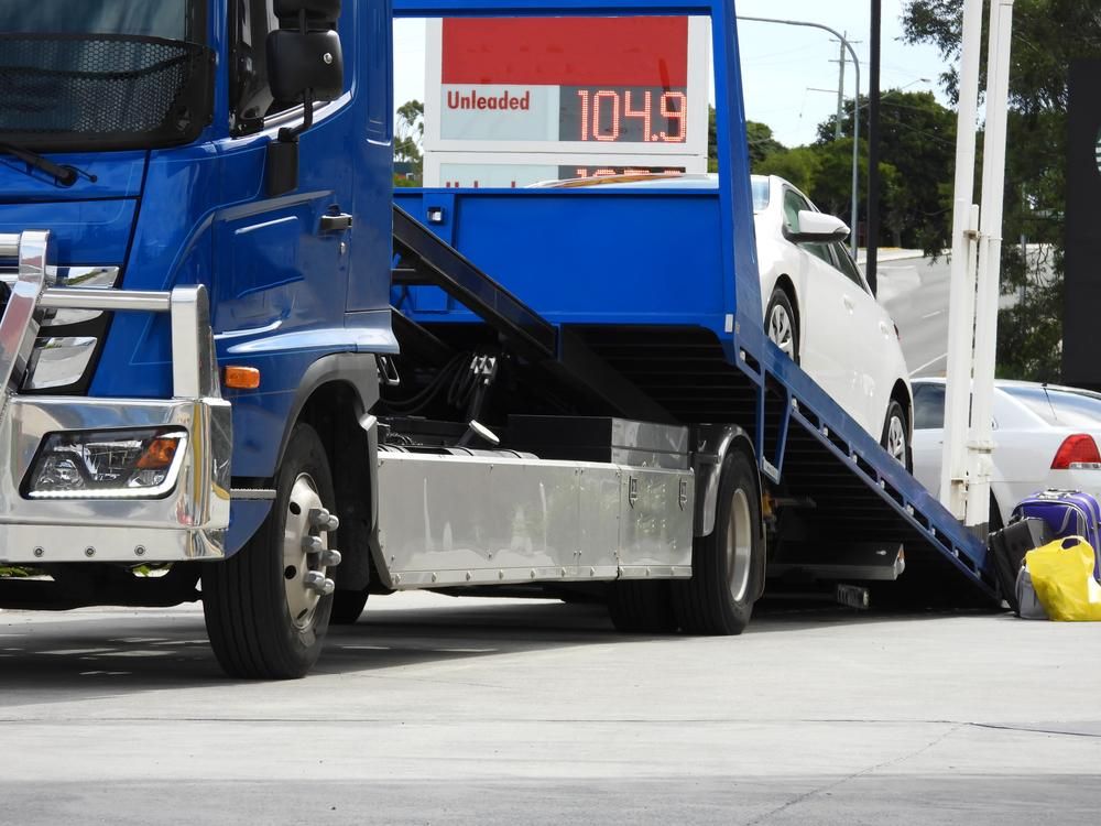 A Blue Tow Truck is Carrying a White Car — DJ's Tilt Tray Service in Coffs Harbour, NSW
