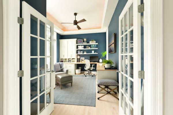 A home office viewed through white French doors, featuring deep blue walls, a desk, shelving, and a gray area rug.