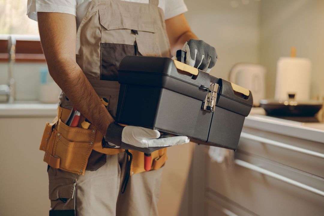 A worker in overalls and work gloves holding a black plastic toolbox in a kitchen.