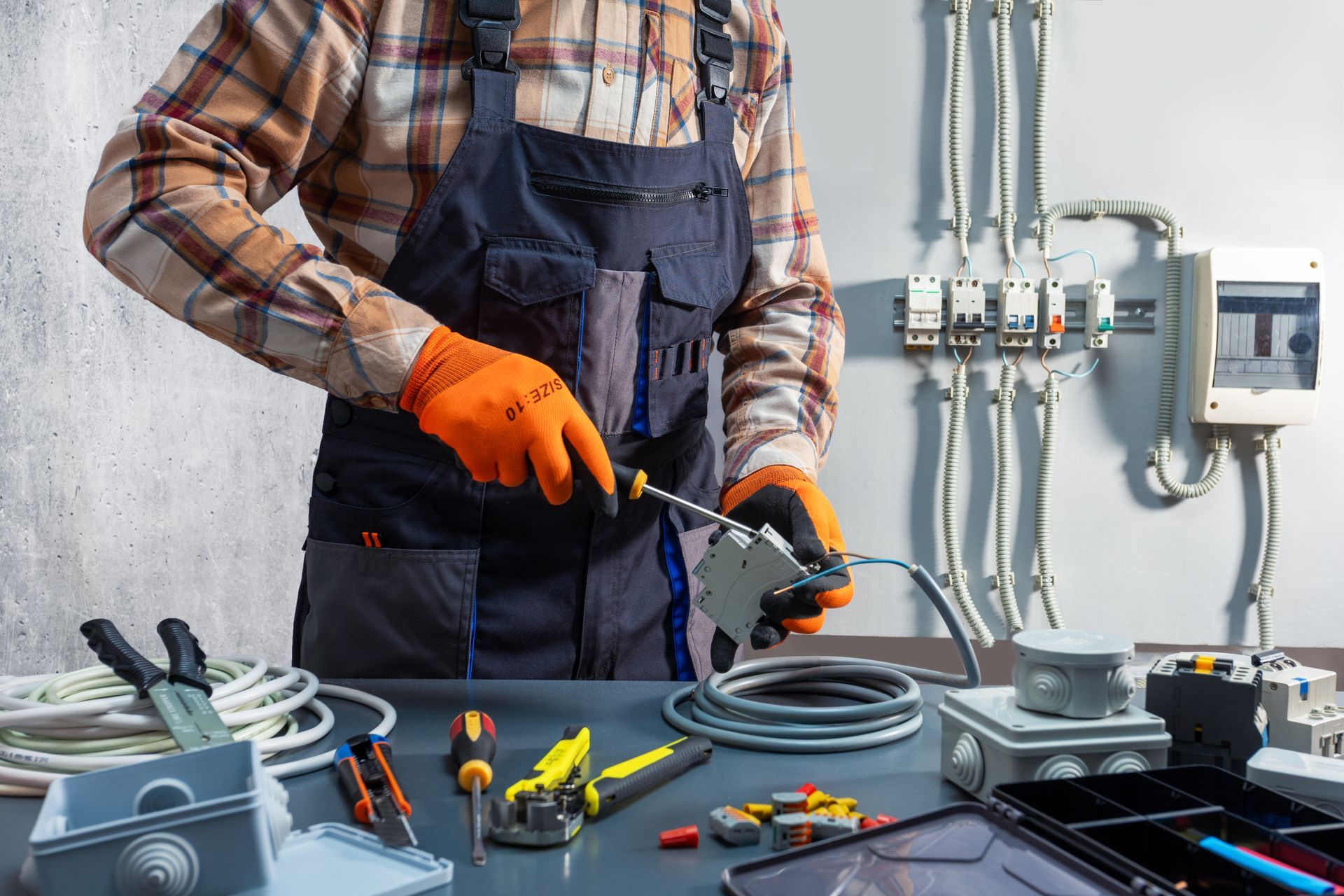 Electrician in orange gloves wiring cables at a workbench with tools and electrical panels nearby
