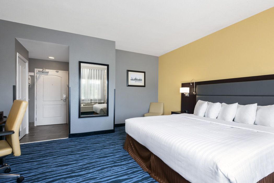 A hotel room featuring a large bed with white linens, blue patterned carpet, a desk, and a wall-mounted mirror.