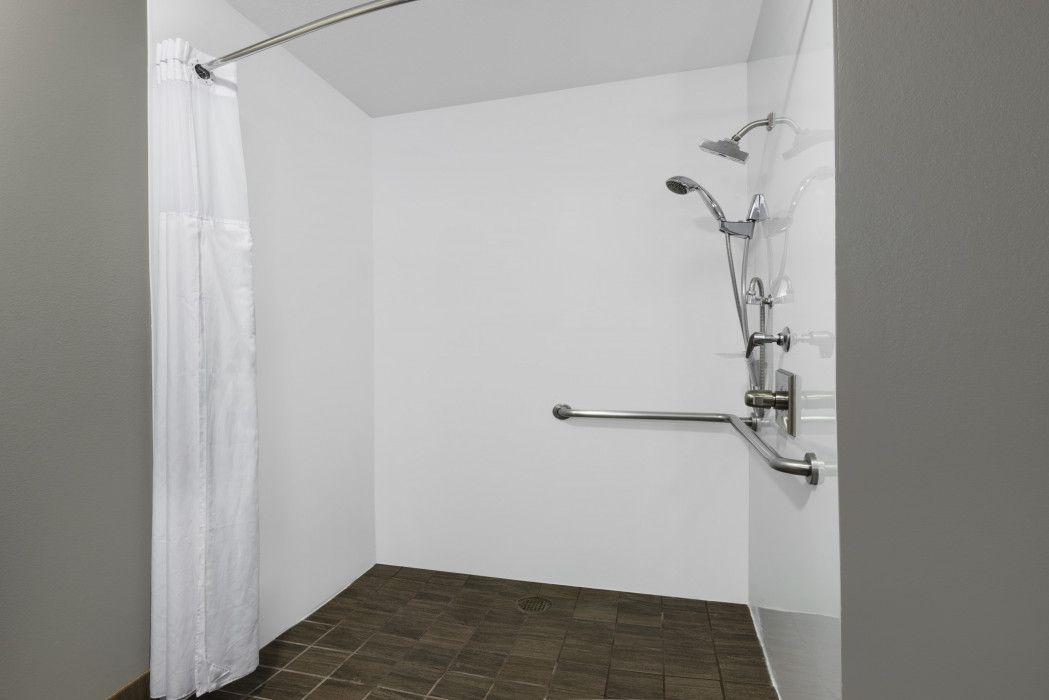 Accessible walk-in shower with white walls, a dark tiled floor, shower head, handheld sprayer, and metal safety grab bars.