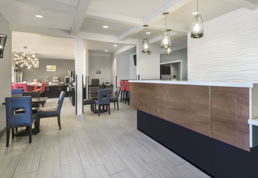 A bright hotel lobby featuring a wooden reception desk, grey tiled floors, and dining tables with blue and red chairs.